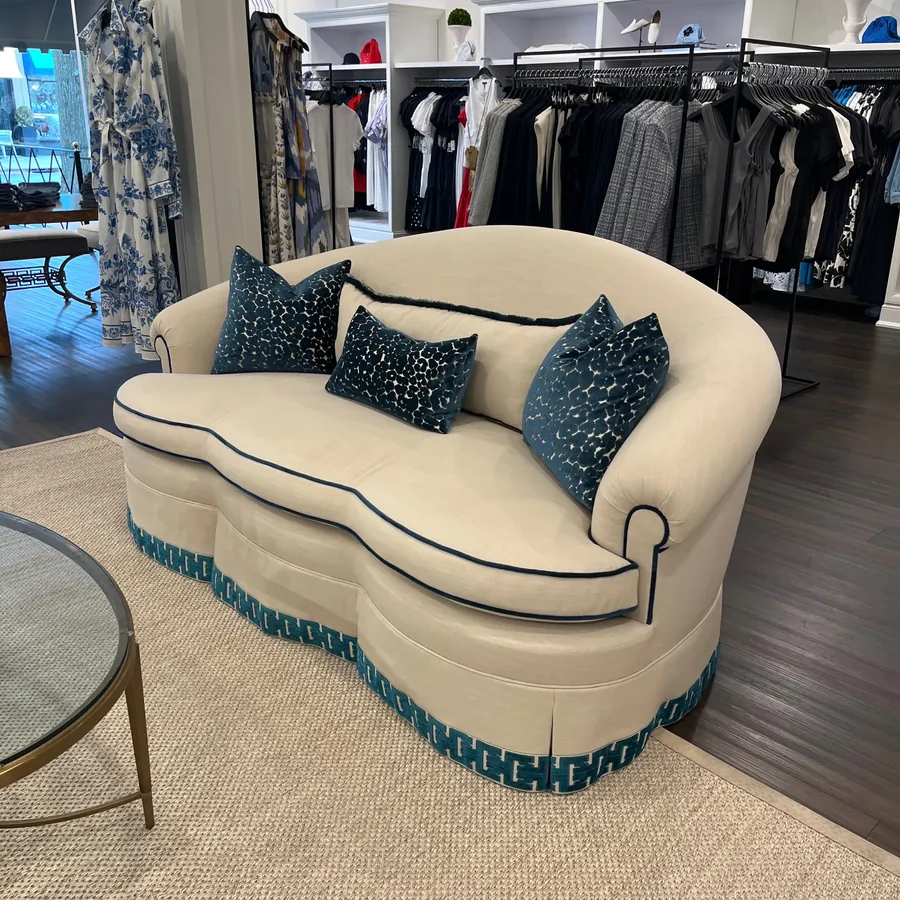 A cream curved sofa with teal pillows and trim beside a glass table