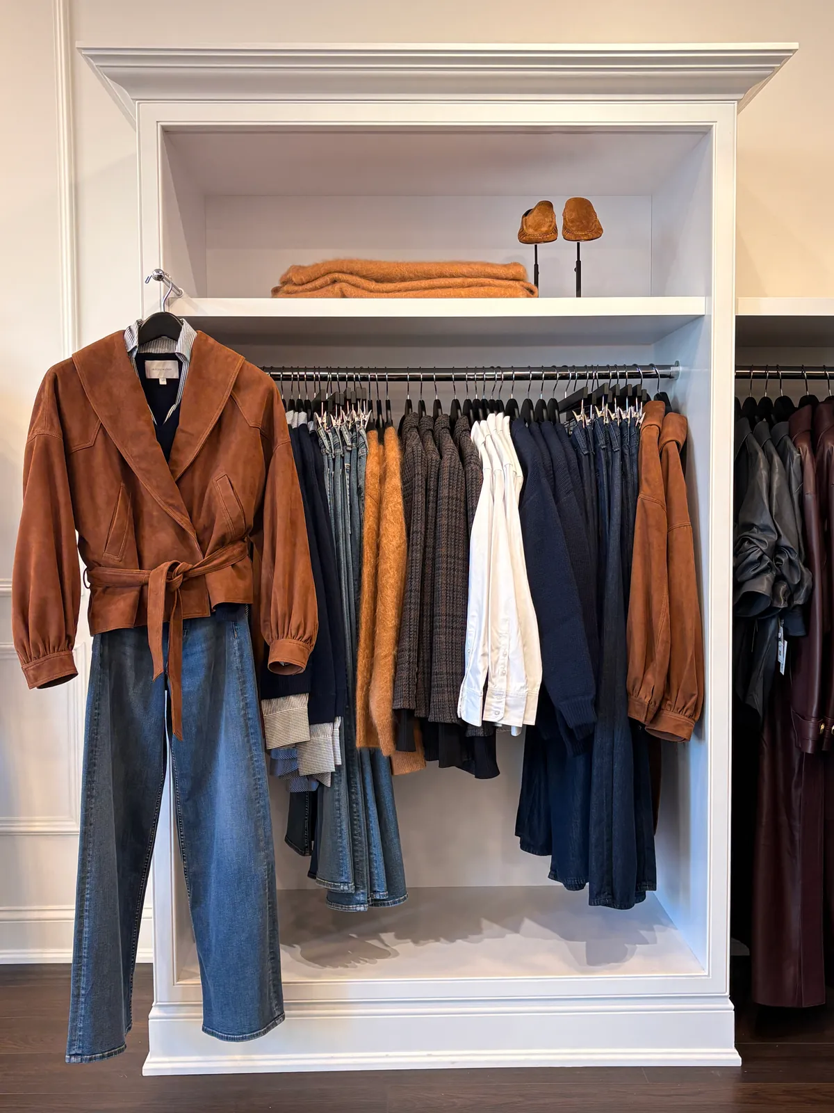 A belted tan suede jacket and flared jeans hung in a white shelving bay, a rail of jackets and shirts behind