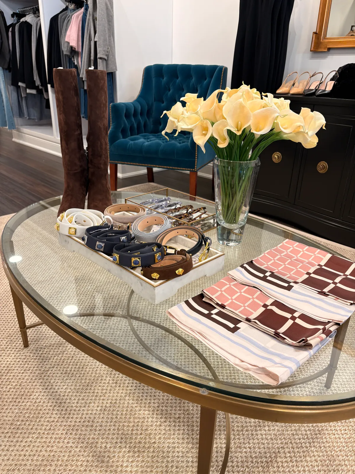 Belts, silk scarves and suede boots on a glass table with yellow calla lilies, a teal velvet chair behind