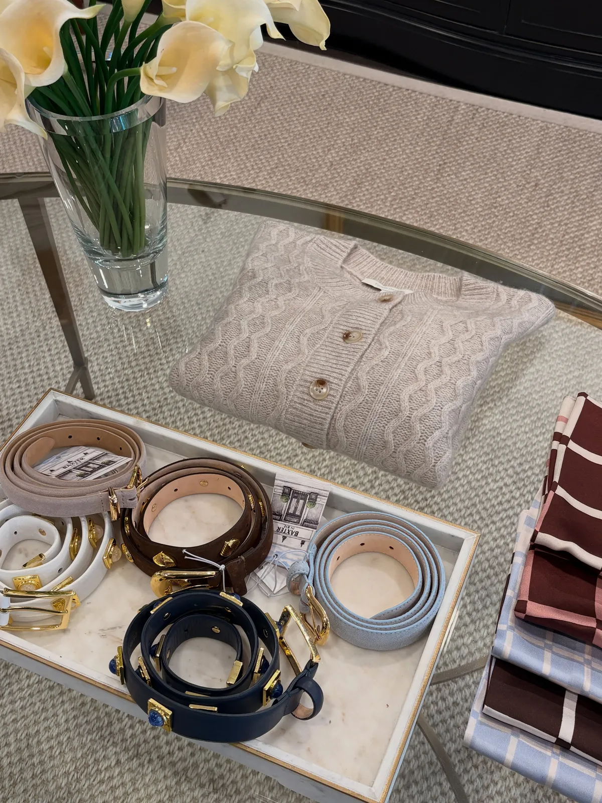 A tray of leather belts in tan, navy and white beside a cable-knit sweater on the glass table