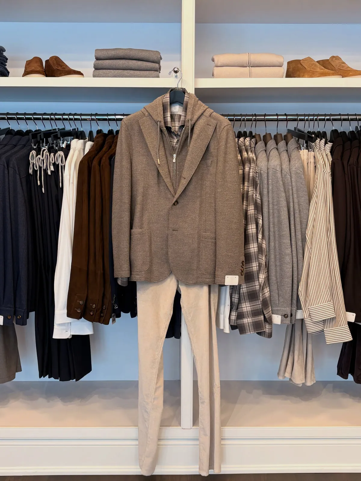 A men’s look on a hook: a brown wool jacket over cream trousers, in front of a rail of jackets and knits with shoes on the shelf above