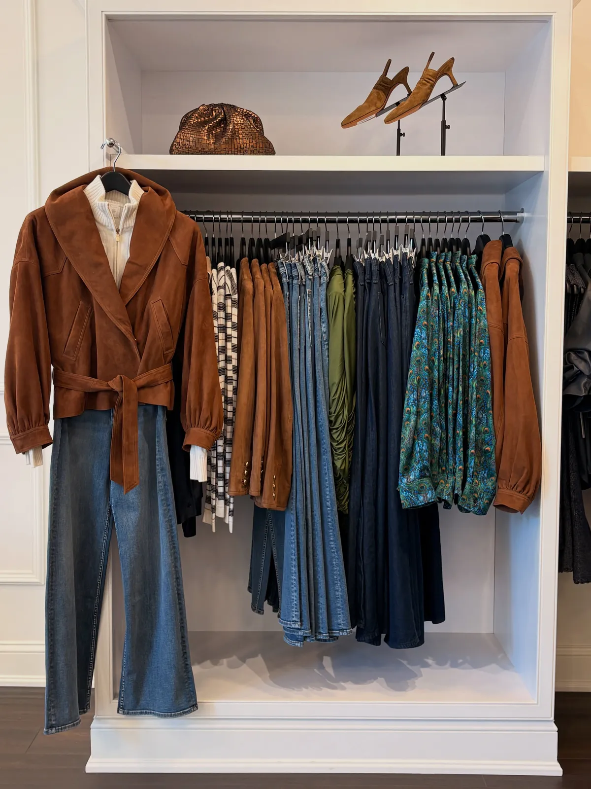 A tan suede jacket with a belted waist styled over wide-leg jeans in front of a rail of blouses